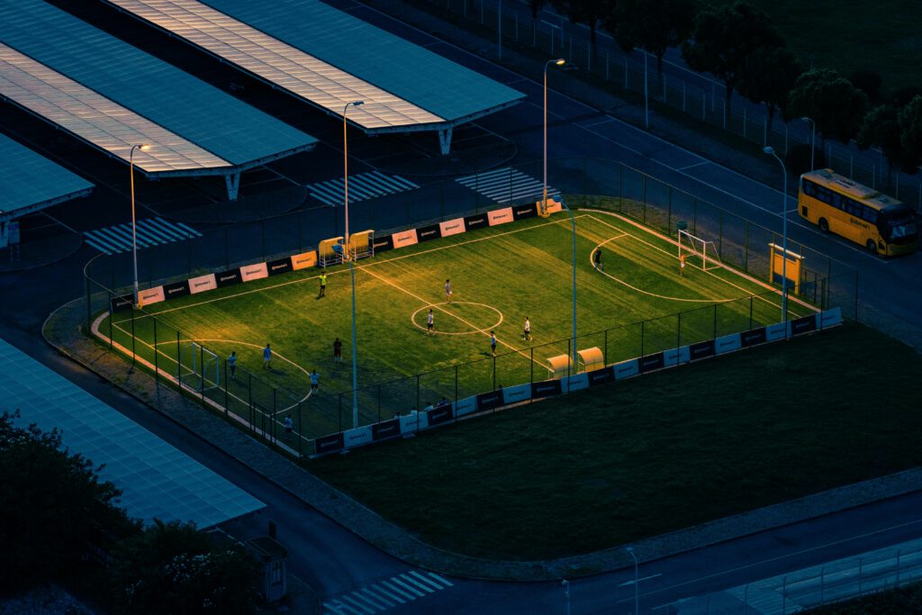pexels-photo-33212390-33212390 Aerial shot of a soccer field lit at night with players, surrounded by urban structures.